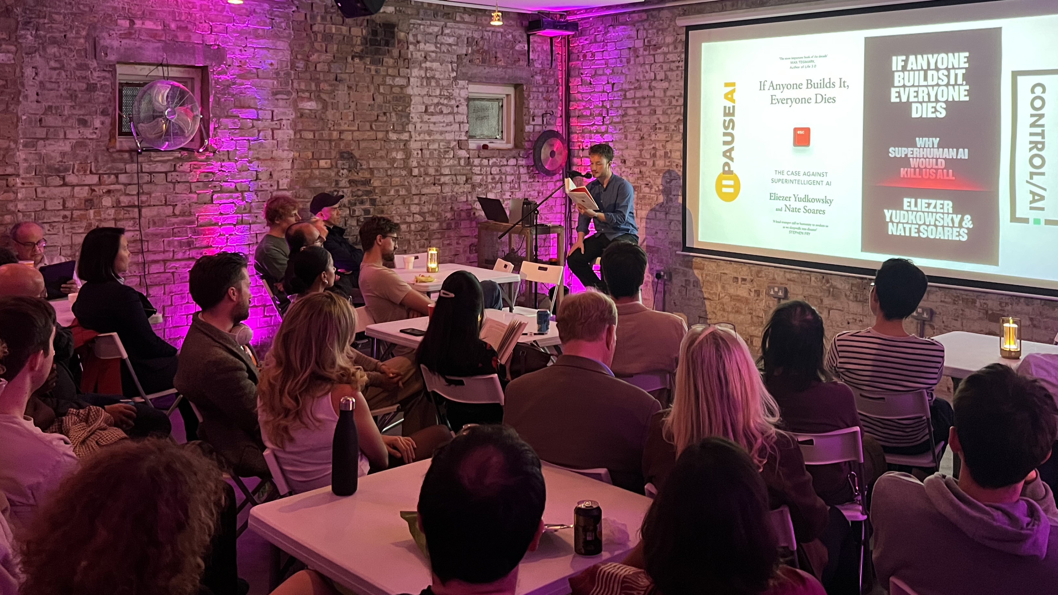 A speaker reads from If Anyone Builds It, Everyone Dies to an audience in a brick-walled room with purple lighting.